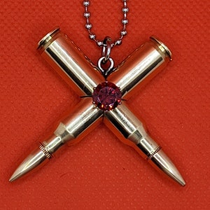 May include: A gold-toned necklace with a pendant made from two bullet casings crossed over each other. A red gemstone is set in the center of the cross.