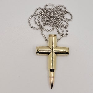 May include: A gold-colored bullet casing cross pendant on a silver chain. The cross is made from four bullet casings, with the bullet tip of one casing forming the bottom point of the cross.