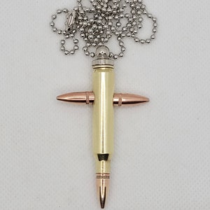 May include: A silver chain necklace with a gold bullet casing cross pendant. The cross is made from a bullet casing with two smaller bullet casings forming the cross arms. The bullet casings are a brass color.
