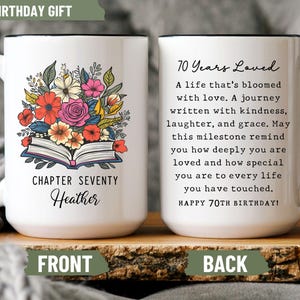 May include: White ceramic mug with a black handle and rim, featuring a floral design with an open book. The front reads "Chapter Seventy Heather" and the back says "70 Years Loved" with a birthday message.