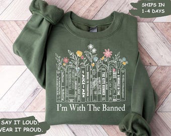 May include: Green sweatshirt with a white graphic of a bookshelf filled with banned books and flowers. The text "I'm With The Banned" is printed below the graphic.