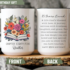 May include: White ceramic mug with a black handle and rim. The front features a floral design above an open book, with the text "CHAPTER EIGHTY-FIVE Heather". The back has text that reads "85 Years Loved" and a birthday message.