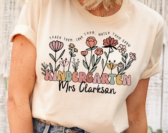 May include: A cream colored t-shirt with a floral design and the text "Teach them, love them, watch them grow. Kindergarten Mrs. Clarkson"