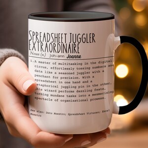 May include: White ceramic mug with a black handle. The mug has a black border around the top and a black and white design that reads "Spreadsheet Juggler Extraordinaire Noun (n.) joh-ann Joanne 1. A master of multitasking in the digital circus, effortlessly tossing numbers and data like a seasoned juggler with a penchant for precision. With a spreadsheet in one hand and a metaphorical juggling pin in the other, this wizard performs dazzling feats, turning mundane tasks into a mesmerizing spectacle of organizational prowess. See also: Data Maestro; Spreadsheet Virtuoso; Excel Maestro"