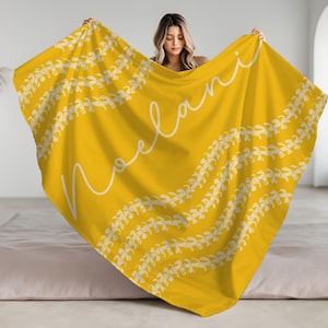 May include: A yellow throw blanket with a white floral pattern and the name "Noelani" written in white cursive script.