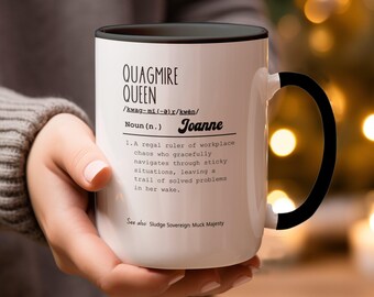 Könnte beinhalten: Eine weiße Keramiktasse mit schwarzem Henkel und Rand. Die Tasse zeigt eine Definition von "Quagmire Queen" mit dem Namen "Joanne" aufgedruckt. Die Definition lautet: "Eine königliche Herrscherin des Chaos am Arbeitsplatz, die mit Anmut durch schwierige Situationen navigiert und eine Spur gelöster Probleme hinterlässt."