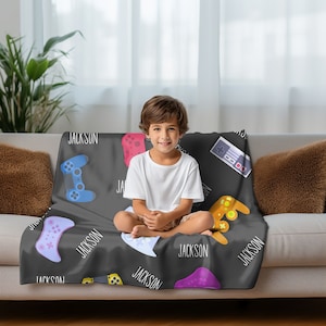 May include: A black fleece blanket with a repeating pattern of colorful video game controllers and the name "Jackson" printed in white.