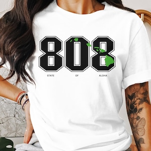 Könnte beinhalten: Weißes T-Shirt mit einer schwarz-weißen Grafik der Zahl 808 mit einem grünen Umriss der hawaiianischen Inseln. Der Text "State of Aloha" befindet sich unter der Grafik.
