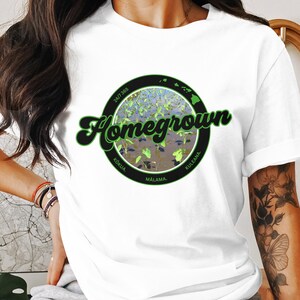 May include: White t-shirt with a black circle graphic featuring a green and blue image of the Hawaiian islands with the text "Homegrown" in a green and white retro font. The text "24/7 365" is in the top left corner of the circle. The text "KOKU'A", "MALAMA", and "KULEANA" are in the bottom of the circle.