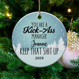 May include: A round, light blue Christmas ornament with the text "YOU ARE A Kick-Ass MANAGER Joanne KEEP THAT SHIT UP 2024". The ornament has a snowy forest design and is hanging on a Christmas tree.