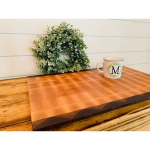 May include: A large, rectangular cutting board made of light wood with a dark wood border. The board has a checkered pattern and is sitting on a wooden surface. A white mug with a green wreath and the letter "M" is on the board.
