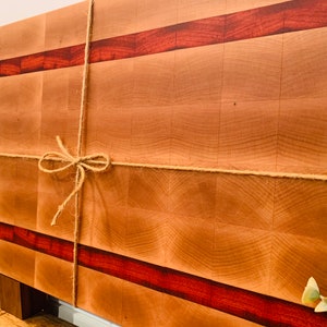 May include: A large, rectangular wooden cutting board with a checkered pattern and two red wood strips. The board is tied with twine and has a wooden stand.