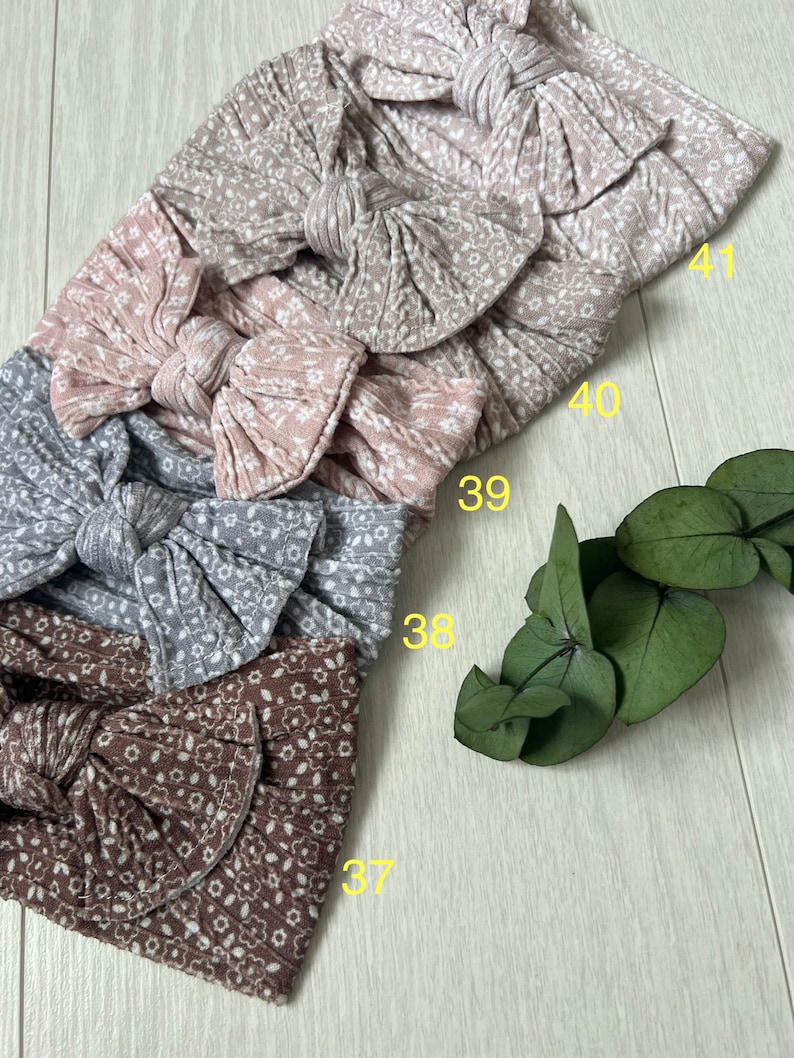 Op de afbeelding: Vier stoffen hoofdbanden met een bloemenpatroon en een strik. De hoofdbanden zijn in de kleuren bruin, grijs, roze en beige. De hoofdbanden zijn genummerd 37, 38, 39, 40 en 41.