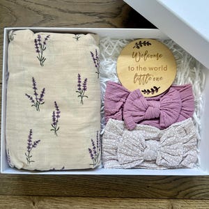 Puede incluir: Una caja de regalo blanca que contiene una manta envolvente beige con ramitas de lavanda moradas, dos diademas de lazo malva y un disco de madera con el texto "Welcome to the world little one."