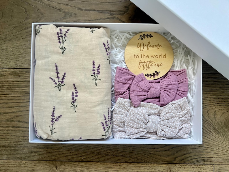 Puede incluir: Una caja de regalo blanca que contiene una manta envolvente beige con ramitas de lavanda moradas, dos diademas de lazo malva y un disco de madera con el texto "Welcome to the world little one."