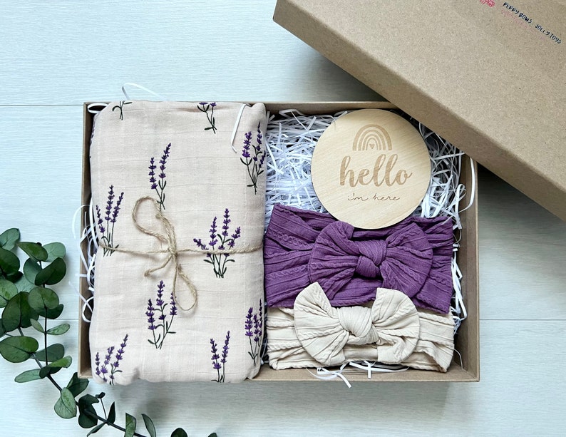 Puede incluir: Una caja de regalo que contiene una manta envolvente de muselina beige con ramitas de lavanda moradas, dos diademas y un disco de madera con el texto "hello i'm here". Las diademas son moradas y beige. La caja es marr&oacute;n.