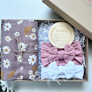 Puede incluir: Una caja de regalo que contiene una manta envolvente de muselina malva con estampado de margaritas blancas y hojas doradas, dos diademas y un disco de anuncio de madera. Las diademas son rosas y blancas. El disco dice "I'm here".