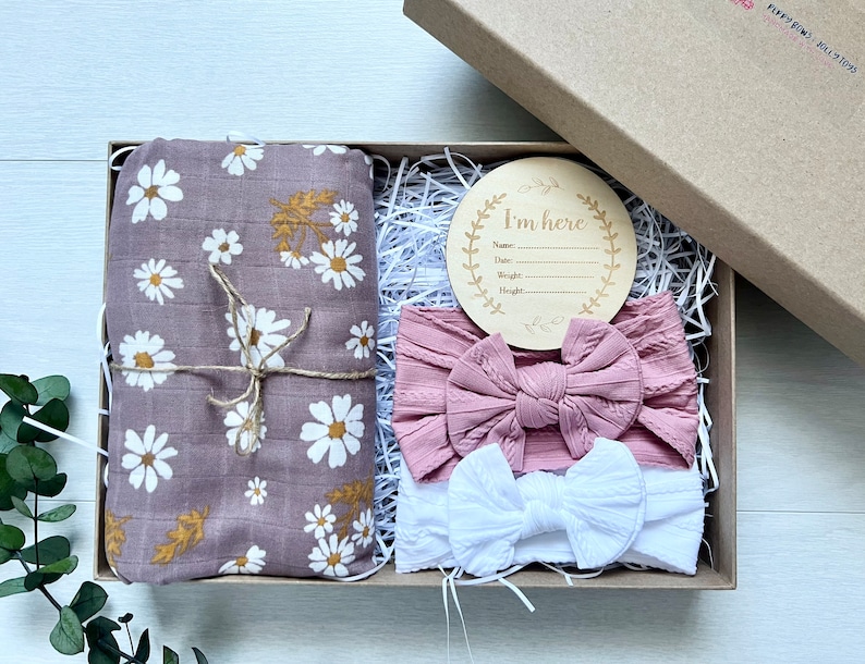 Puede incluir: Una caja de regalo que contiene una manta envolvente de muselina malva con estampado de margaritas blancas y hojas doradas, dos diademas y un disco de anuncio de madera. Las diademas son rosas y blancas. El disco dice "I'm here".