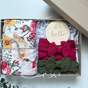 Puede incluir: Una caja de regalo con una manta floral, dos diademas y un disco de madera. La manta tiene flores amarillas, rosas y rojas. Las diademas son burdeos y verde oliva. El disco de madera dice "hello I'm here".