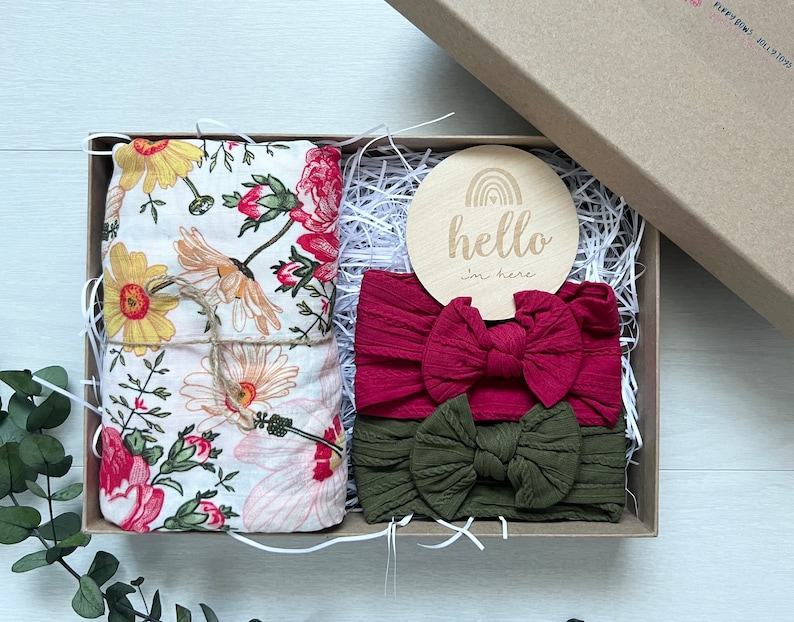 Puede incluir: Una caja de regalo con una manta floral, dos diademas y un disco de madera. La manta tiene flores amarillas, rosas y rojas. Las diademas son burdeos y verde oliva. El disco de madera dice "hello I'm here".