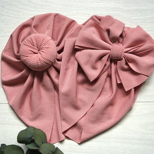 May include: A pink ribbed fabric turban with a large bow. The turban is folded into a heart shape.