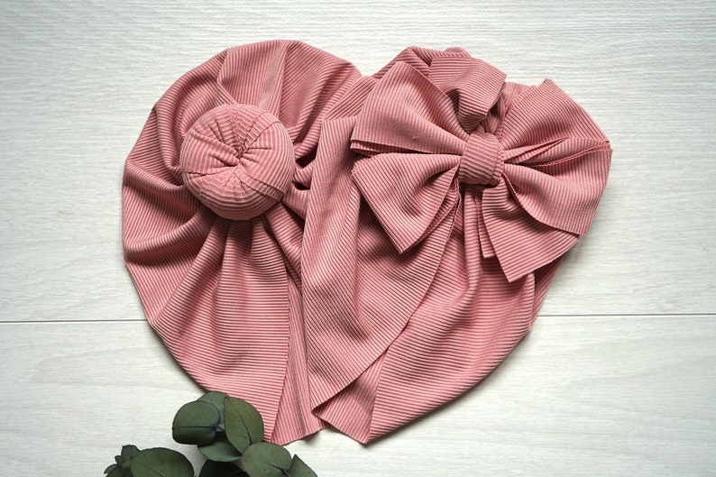 May include: A pink ribbed fabric turban with a large bow. The turban is folded into a heart shape.