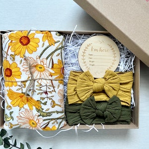 Puede incluir: Caja de regalo con una muselina con estampado de margaritas amarillas, dos diademas anudadas en amarillo mostaza y verde oliva, y un disco de anuncio de madera con la inscripci&oacute;n "I'm here". La caja est&aacute; rellena de papel.