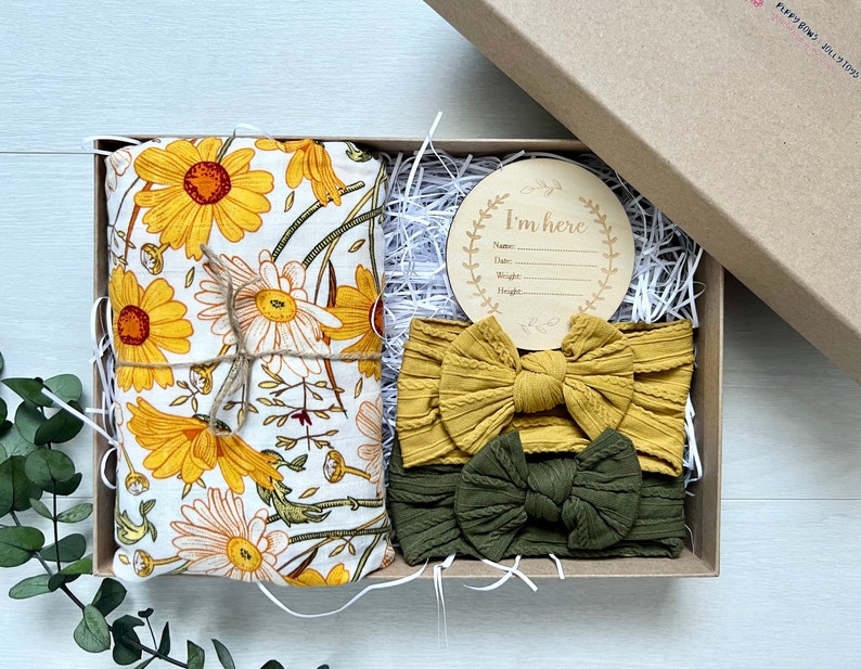 Puede incluir: Caja de regalo con una muselina con estampado de margaritas amarillas, dos diademas anudadas en amarillo mostaza y verde oliva, y un disco de anuncio de madera con la inscripci&oacute;n "I'm here". La caja est&aacute; rellena de papel.