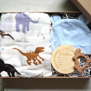 Puede incluir: Un set de regalo para beb&eacute; en una caja marr&oacute;n, que incluye una muselina blanca con ilustraciones de dinosaurios, un gorro azul claro con orejas de oso, un juguete de dentici&oacute;n de madera y una placa de anuncio de madera que dice "I'm here".