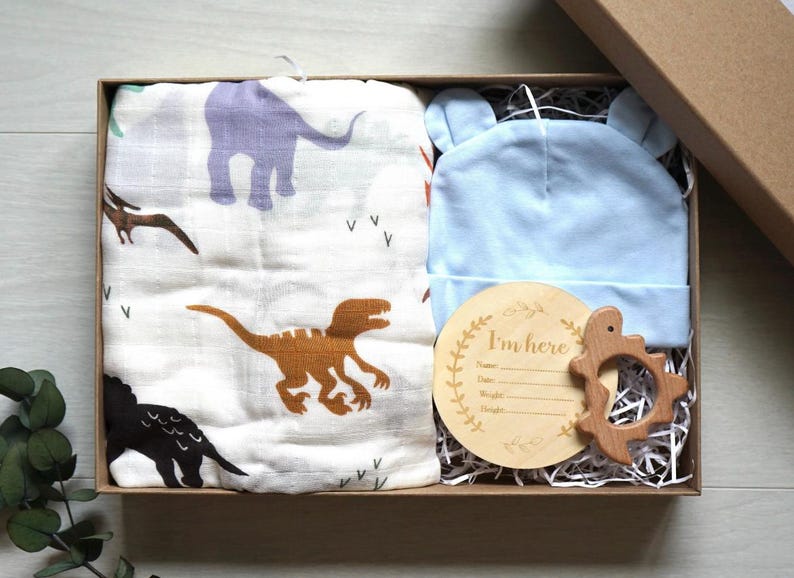 Puede incluir: Un set de regalo para beb&eacute; en una caja marr&oacute;n, que incluye una muselina blanca con ilustraciones de dinosaurios, un gorro azul claro con orejas de oso, un juguete de dentici&oacute;n de madera y una placa de anuncio de madera que dice "I'm here".