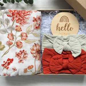 Puede incluir: Caja de regalo con una muselina floral, dos diademas de punto trenzado con lazos y un disco de madera con "hello I'm here" y un arco&iacute;ris. Los art&iacute;culos son de color naranja, crema y beige. La caja est&aacute; llena de papel.