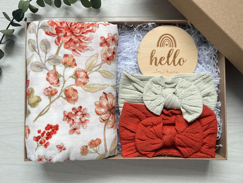 Puede incluir: Caja de regalo con una muselina floral, dos diademas de punto trenzado con lazos y un disco de madera con "hello I'm here" y un arco&iacute;ris. Los art&iacute;culos son de color naranja, crema y beige. La caja est&aacute; llena de papel.