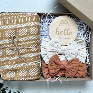 Puede incluir: Una caja de regalo que contiene una manta estampada enrollada y atada con hilo, dos diademas con lazo y un disco de madera con el texto "hello I'm here" y un dise&ntilde;o de arco&iacute;ris. La manta es de color beige con patrones geom&eacute;tricos blancos.