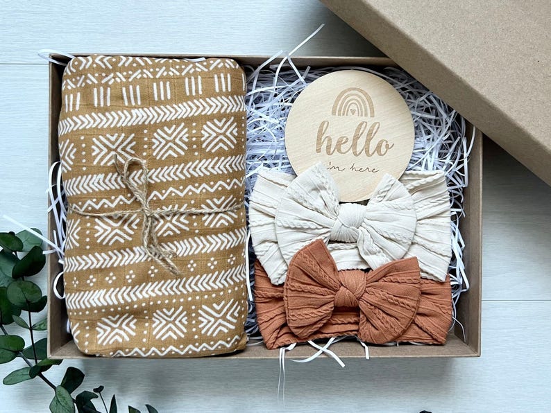 Puede incluir: Una caja de regalo que contiene una manta estampada enrollada y atada con hilo, dos diademas con lazo y un disco de madera con el texto "hello I'm here" y un dise&ntilde;o de arco&iacute;ris. La manta es de color beige con patrones geom&eacute;tricos blancos.