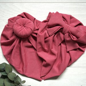 May include: A pink stretchy fabric headband with a large bow and a bun maker. The headband is made of a ribbed fabric and is perfect for adding a touch of style to any outfit.