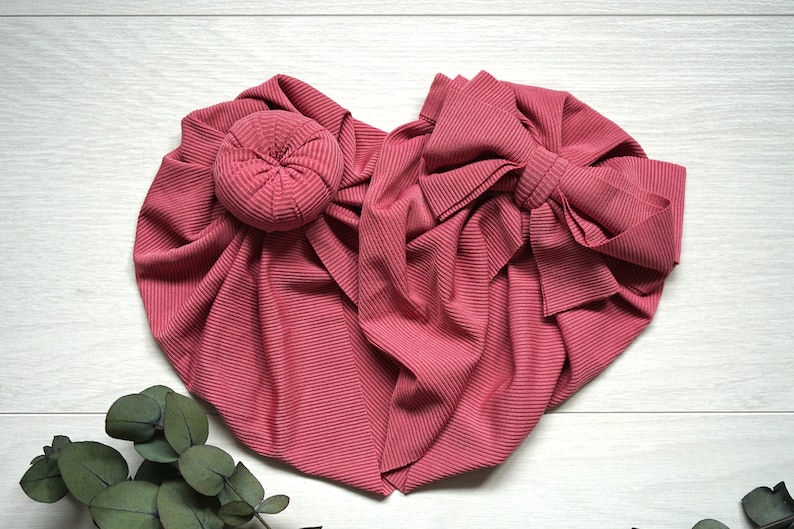 May include: A pink stretchy fabric headband with a large bow and a bun maker. The headband is made of a ribbed fabric and is perfect for adding a touch of style to any outfit.