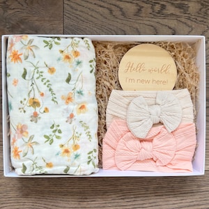 Puede incluir: Una caja de regalo blanca contiene una muselina floral para envolver, un disco de madera con el texto "Hello world. I'm new here!" y dos diademas para beb&eacute;. Las diademas son crema y rosa claro con detalles de lazo.