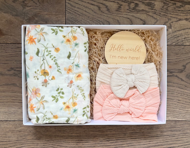 Puede incluir: Una caja de regalo blanca contiene una muselina floral para envolver, un disco de madera con el texto "Hello world. I'm new here!" y dos diademas para beb&eacute;. Las diademas son crema y rosa claro con detalles de lazo.