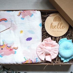 Puede incluir: Un juego de regalo para beb&eacute;s en una caja de cart&oacute;n. El juego incluye una manta envolvente de muselina blanca con ilustraciones de unicornios rosas, una diadema de punto rosa, una diadema de punto azul y un disco de madera con el texto "hello I'm here".