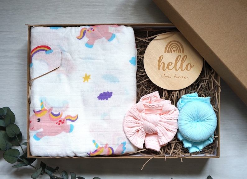 Puede incluir: Un juego de regalo para beb&eacute;s en una caja de cart&oacute;n. El juego incluye una manta envolvente de muselina blanca con ilustraciones de unicornios rosas, una diadema de punto rosa, una diadema de punto azul y un disco de madera con el texto "hello I'm here".