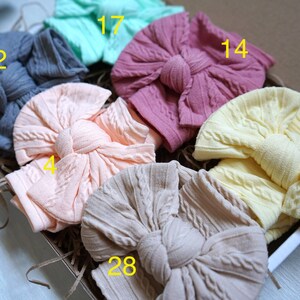 Op de afbeelding: Een set van zes gebreide hoofdbanden in verschillende kleuren, waaronder perzik, lichtroze, beige, geel, grijs en teal. Elke hoofdband heeft een grote, geknoopte strik.