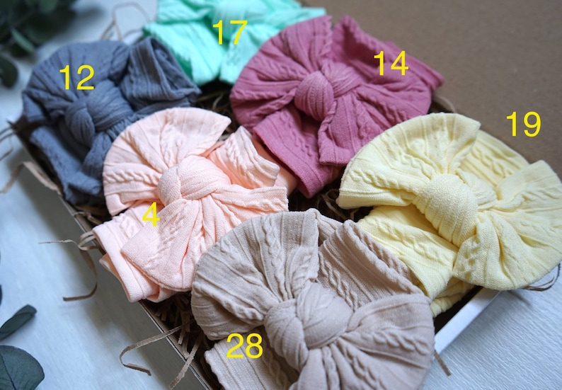 Op de afbeelding: Een set van zes gebreide hoofdbanden in verschillende kleuren, waaronder perzik, lichtroze, beige, geel, grijs en teal. Elke hoofdband heeft een grote, geknoopte strik.