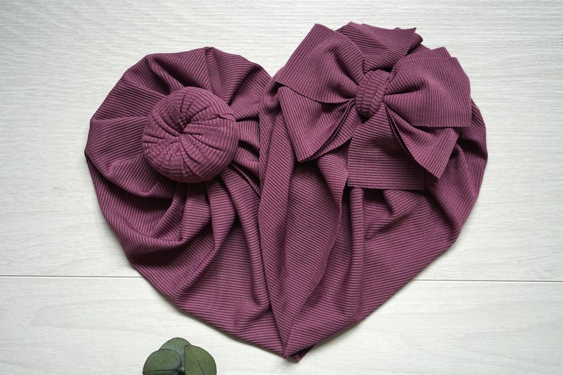 May include: Two mauve fabric headbands, one with a large bow and the other with a bun. The headbands are made of a ribbed fabric.