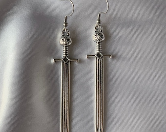May include: A pair of silver sword-shaped dangle earrings. The swords have a detailed design and a small crown at the top.
