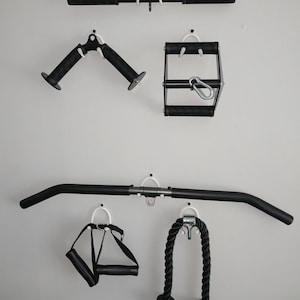 May include: A collection of black gym cable attachments mounted on a wall, including a straight bar, a V-shaped handle, a rectangular handle with a carabiner, a long lat pulldown bar, a single strap handle, and a thick black tricep rope attachment.