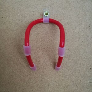 K&ouml;nnte beinhalten: Ein roter, U-f&ouml;rmiger Werkzeughalter mit rosa Gummimanschetten an den Enden und in der Mitte. Der Halter ist auf einer hellbraunen Oberfl&auml;che mit einem transparenten Kunststoffclip und einer Schraube montiert.