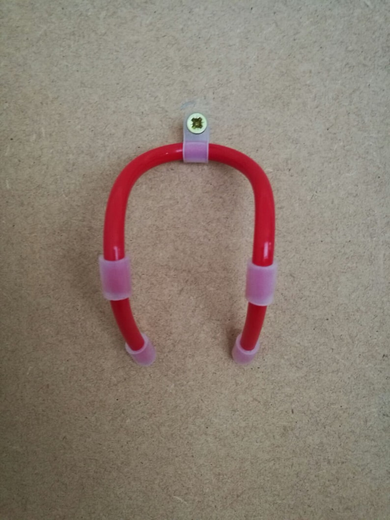K&ouml;nnte beinhalten: Ein roter, U-f&ouml;rmiger Werkzeughalter mit rosa Gummimanschetten an den Enden und in der Mitte. Der Halter ist auf einer hellbraunen Oberfl&auml;che mit einem transparenten Kunststoffclip und einer Schraube montiert.