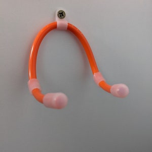 K&ouml;nnte beinhalten: Ein orangefarbener, flexibler U-f&ouml;rmiger Kleiderb&uuml;gel mit rosa Endkappen und Halterungen. Der Kleiderb&uuml;gel ist mit einer Schraube an einer wei&szlig;en Oberfl&auml;che befestigt. Das Design ist einfach und funktional.