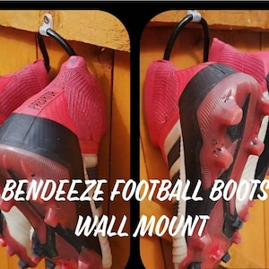 Puede incluir: Dos pares de botas de fútbol rojas y negras colgadas de un soporte de pared. Las botas están etiquetadas con "Predator". El texto "Bendeezee Football Boots Wall Mount" está escrito debajo de las botas.
