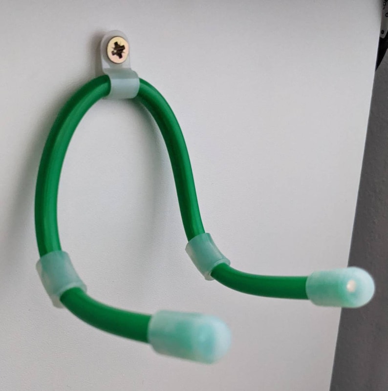 K&ouml;nnte beinhalten: Ein gr&uuml;nes, flexibles, U-f&ouml;rmiges Ger&auml;t mit hellgr&uuml;nen Endkappen und Befestigungsmaterial. Das Ger&auml;t ist mit einer Schraube an einer wei&szlig;en Oberfl&auml;che befestigt. Das flexible Design erm&ouml;glicht die Anpassung und Positionierung.